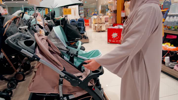مفهوم تجهيز مستلزمات المولود الجديد، تصوير من الخلف لامرأة عربية خليجية اماراتية محجبة تقوم باختيار عربة أطفال داخل متجر مستلزمات الأطفال، التسوق داخل المولات التجارية الحديثة