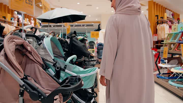 اختيار مستلزمات الأطفال بجودة عالية، تصوير من الخلف لامرأة عربية خليجية اماراتية محجبة تقوم باختيار عربة أطفال داخل متجر مستلزمات الأطفال، مفهوم تجهيز مستلزمات المولود الجديد، التسوق داخل المولات التجارية الحديثة