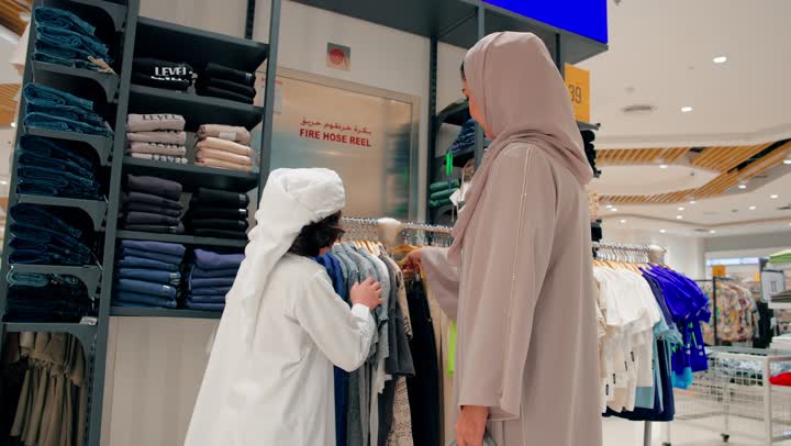 الاهتمام بالمظهر والهوية الخليجية، اختيار ثوب مناسب للمقاس، ولدعربي خليجي يرتدي الزي التقليدي يقوم بتجربة ثوب أطفال جديد داخل متجر ملابس برفقة والدته، التسوق العائلي داخل المتاجر التجارية الحديثة