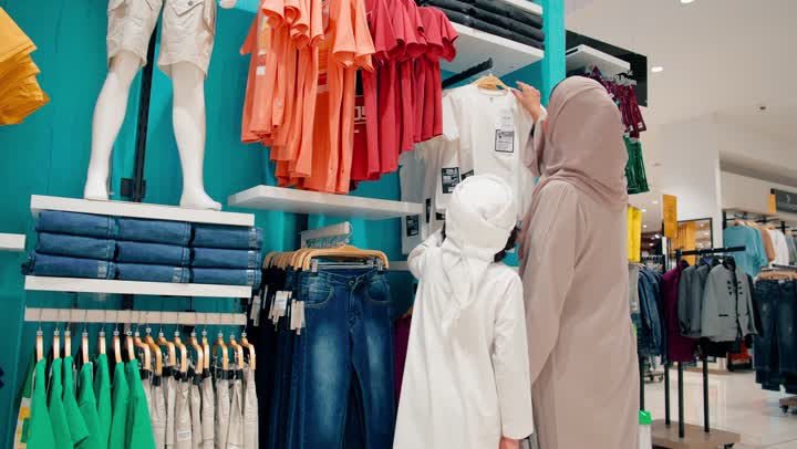 اختيار ثوب مناسب للمقاس، ولدعربي خليجي يرتدي الزي التقليدي يقوم بتجربة ثوب أطفال جديد داخل متجر ملابس برفقة والدته، التسوق العائلي داخل المتاجر التجارية الحديثة، الاهتمام بالمظهر والهوية الخليجية