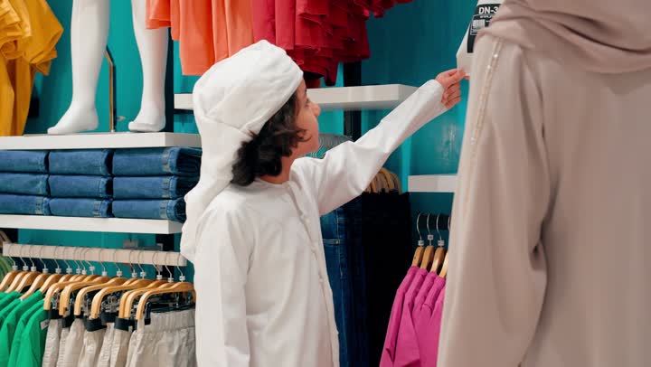 A Gulf Arab boy wearing traditional attire is trying on a new children's outfit inside a clothing store with his mother, choosing a suitable size, family shopping in modern retail stores, and paying attention to appearance and Gulf identity.