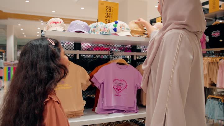 بنت عربيةخليجية امارتية تقوم بتجربة قبعة أطفال داخل متجر ملابس وإكسسوارات برفقة والدتها، مفهوم التسوق العائلي داخل المولات التجارية، تجربة تسوق ممتعة للأطفال، الاهتمام بمظهر الأطفال وأناقتهم