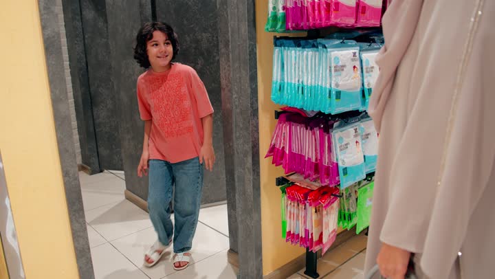 The relationship of motherhood and caring for children while shopping, an Emirati Arab boy trying on a shirt inside a clothing store with his mother, the concept of family shopping in commercial stores, carefully and thoughtfully choosing children's clothes, and paying attention to children's appearance and elegance.