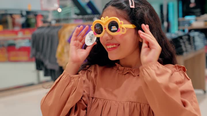 تصوير مقرب لبنت عربيه خليجيه اماراتية ترتدي الزي الكاجوال تقوم بتجربة نظارة شمسية ملونة داخل متجر ملابس وإكسسوارات،ايماءات السعادة و السرور ، مفهوم التسوق العائلي داخل المولات التجارية الحديثة، الاهتمام بمظهر الأطفال وأناقتهم
