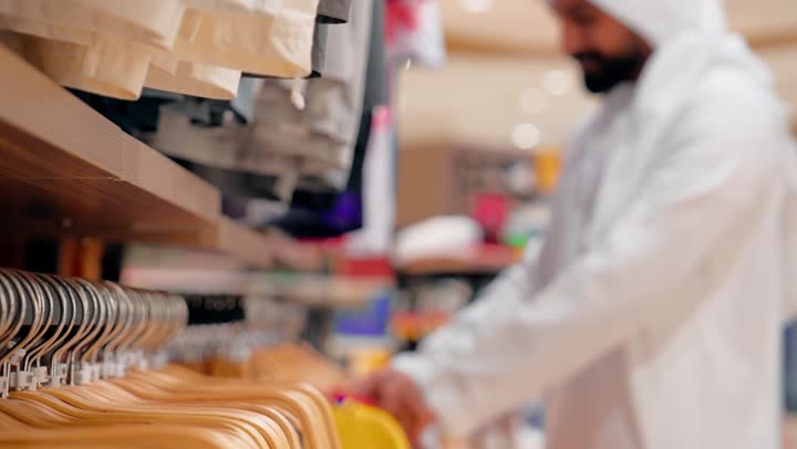 الموضة الرجالية في المجتمعات الخليجية، مفهوم التسوق العصري في المولات الحديثة، اختيار الملابس المناسبة بعناية، تصوير من الجانب لرجل عربي خليجي يرتدي الزي التقليدي الإماراتي يقوم باختيار قميص رجالي عصري، الأناقة الرجالية والاهتمام بالمظهر الخارجي