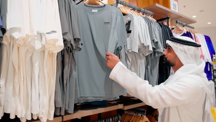 مفهوم التسوق العصري في المولات الحديثة، اختيار الملابس المناسبة بعناية، رجل عربي خليجي يرتدي الزي التقليدي الإماراتي يقوم باختيار قميص رجالي عصري، الأناقة الرجالية والاهتمام بالمظهر الخارجي، الموضة الرجالية في المجتمعات الخليجية