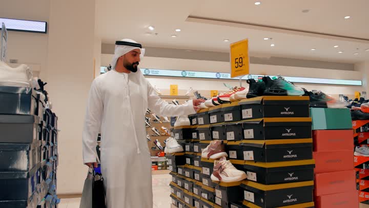 The concept of elegance and quality of choice, shopping inside a modern mall, an Arab Gulf Emirati man wearing a kandura and ghutrah strolling between the sections of shoes and clothing.