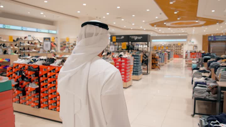 التسوق داخل مول عصري، تصوير من الخلف لرجل عربي خليجي إماراتي يرتدي الكندورة و الغتره يتجول بين أقسام الأحذية والملابس، مفهوم الأناقة وجودة الاختيار