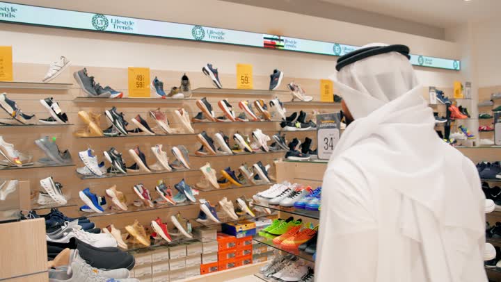 مفهوم الأناقة والراحة، التسوق داخل متجر أحذية، تصوير من الخلف لرجل عربي خليجي إماراتي يرتدي الكندورة و الغترة يتفقد الأحذية الرجالية المعروضة على الأرفف، اختيار الحذاء المناسب بعناية