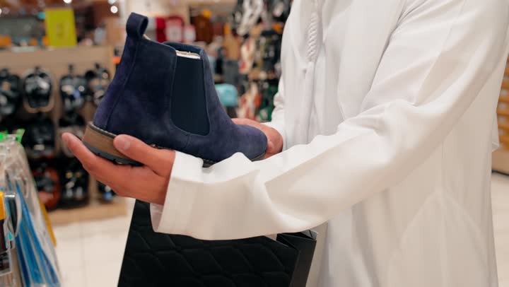 عناية بالاختيار وجودة الخامات، مفهوم الأناقة الرجالية والموضة المحافظة، تصوير مقرب ليد رجل عربي خليجي إماراتي يرتدي الكندورة يتسوق بين تشكيلة متنوعة من الأحذية الرجالية، اختيار الأحذية داخل متجر تجزئة