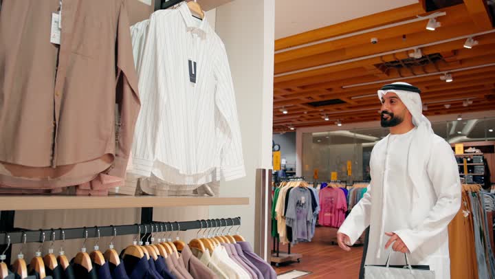 مفهوم التسوق والاهتمام بالمظهرالخارجي، اختيار الأزياء بعناية، التسوق داخل المول، رجل عربي خليجي إماراتي يرتدي الكندورة و الغتره يتفقد القمصان الرجالية المعروضة