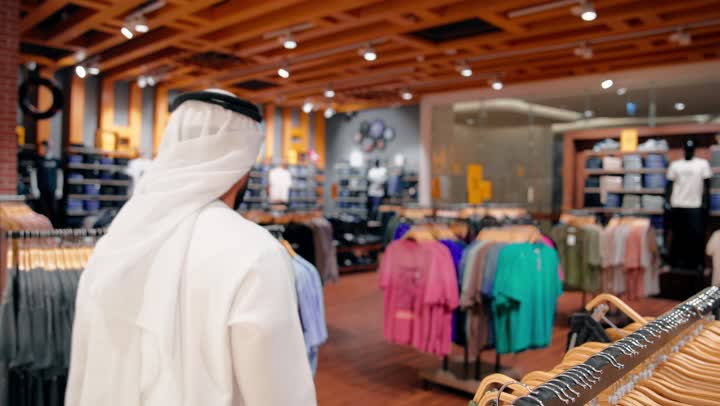 اختيار الأزياء بعناية، التسوق داخل المول،  تصوير من الخلف لرجل عربي خليجي إماراتي يرتدي الكندورة و الغتره يتجول بين أقسام الملابس الجاهزة، مفهوم التسوق والاهتمام بالمظهر