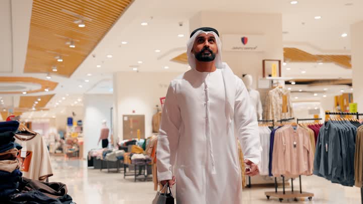 التسوق داخل المول، رجل عربي خليجي إماراتي يرتدي الكندورة و الغتره يتجول بين أقسام الملابس الجاهزة، اختيار الأزياء بعناية، مفهوم التسوق والاهتمام بالمظهر