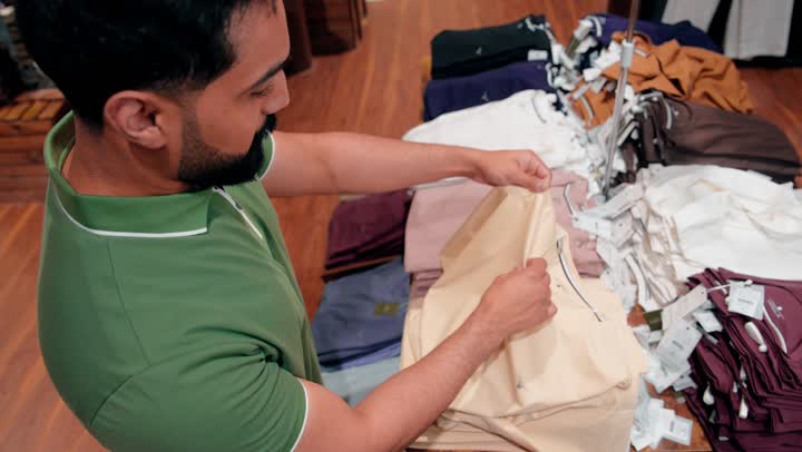 اختيار دقيق للمنتجات قبل الشراء، التسوق داخل متجر ملابس، تصوير مقرب لرجل عربي خليجي اماراتي يرتدي الزي الكاجوال يقوم بتفقد جودة الأقمشة ومقاسات الملابس الجاهزة، مفهوم التسوق الواعي والانتباه للتفاصيل، تجربة شراء داخل متجر أزياء رجالي