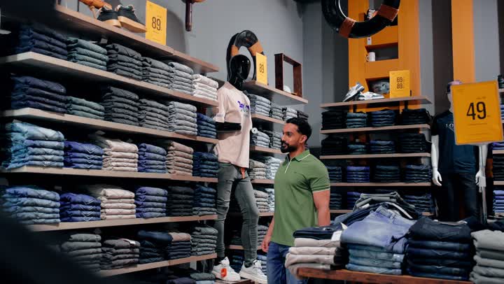 التركيز على جودة الأقمشة وتنوع الخيارات، مفهوم التسوق والاختيار بعناية، شاب عربي خليجي اماراتي  يرتدي الزي الكاجوال يتفحص بنطال جينز مطوي على الرفوف، التسوق داخل متجر ملابس عصري