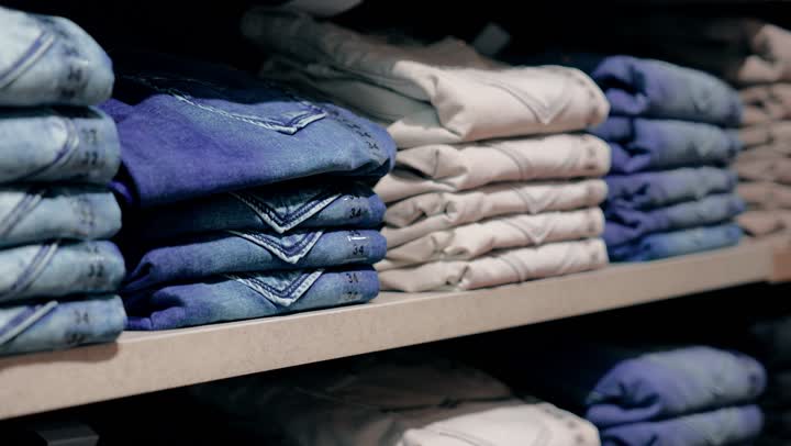 مفهوم التسوق والاختيار بعناية، تصوير مقرب ليد عميل عربي خليجي اماراتي يتفحص بنطال جينز مطوي على الرفوف،  التسوق داخل متجر ملابس عصري، التركيز على جودة الأقمشة وتنوع الخيارات