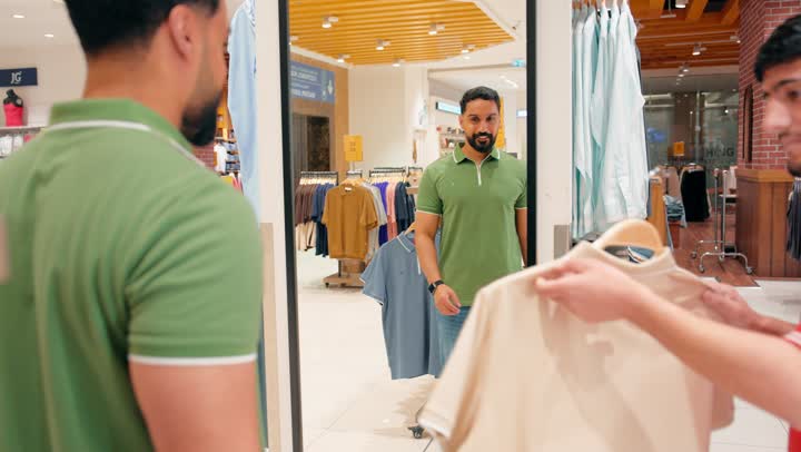 التسوق داخل متجر ملابس ، موظف مبيعات عربي خليجي اماراتي يرتدي اليونيفورم يعرض قميص رجالي على العميل، تفاعل مباشر بين البائع والزبون، اختيار الملابس الجاهزة بعناية، خدمة عملاء داخل متجر تجزئة حديث، أجواء تسوق مريحة ومنظمة