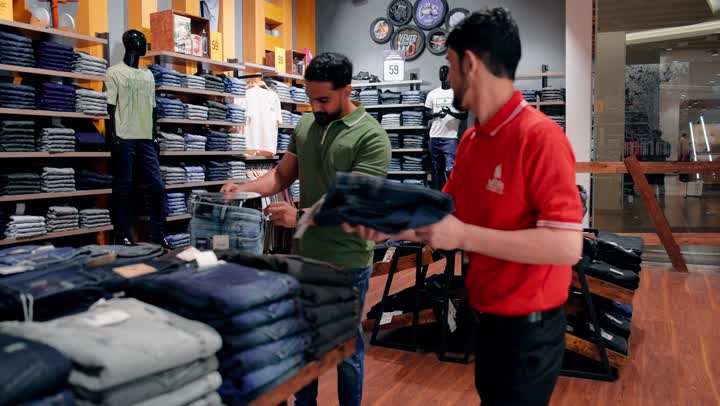 تفاعل مباشر بين البائع والعميل، عرض بنطال جينز ومقارنته بالمقاس المناسب، موظف مبيعات عربي خليجي اماراتي يساعد الزبون داخل متجر ملابس، تجربة تسوق داخل متجر تجزئة حديث، تنظيم وعرض الملابس الجاهزة داخل المحل