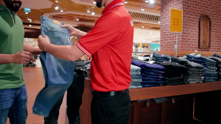 Displaying jeans and comparing them to the appropriate size, an Emirati Gulf Arab sales employee assists the customer inside a clothing store, a shopping experience inside a modern retail store, direct interaction between the seller and the customer, organizing and displaying ready-made clothes inside the shop.