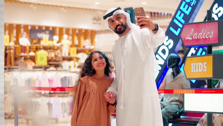 The concept of family bonding and family happiness, an Arab Gulf Emirati father wearing traditional attire takes a selfie with his daughter inside a modern shopping mall.