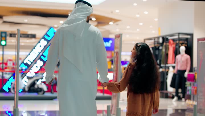 التسوق مع العائلة، تصوير من الخلف لأب عربي خليجي إماراتي يرتدي الكندورة و الغتره يمشي مع ابنته داخل مركز تجاري حديث، مفهوم الترابط الأسري