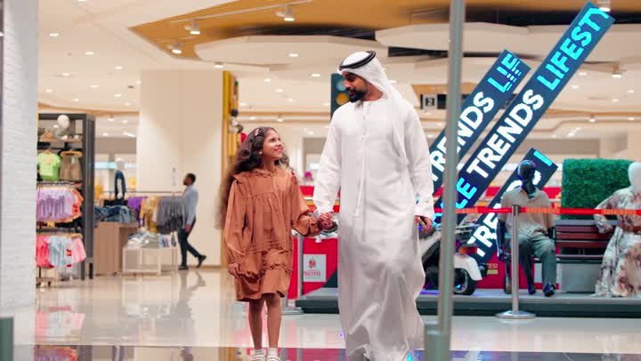 التسوق العائلي داخل مركز تجاري حديث، أب عربي خليجي إماراتي يرتدي الكندورة و الغتره يمشي ممسكا بيد ابنته، مفهوم الترابط الأسري، ايماءات السعادة و السرور