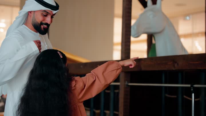 أب عربي خليجي إماراتي  يرتدي الكندورة و الغتره يرافق ابنته داخل مركز تسوق حديث، بنت تشير بيدها نحو مجسم حصان خشبي، مفهوم التفاعل الأسري والفضول الطفولي، قضاء وقت عائلي ممتع داخل مول تجاري