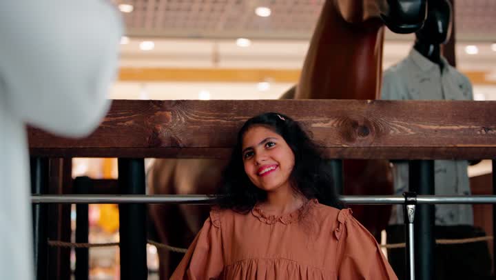 تصوير مقرب لفتاة عربية خليجية إماراتية مبتسمة ترتدي الزي الكاجوال تقف داخل مول تجاري بالقرب من الألعاب الترفيهية، مفهوم السعادة والطفولة والأجواء الترفيهي، ايماءات السعادة و السرور