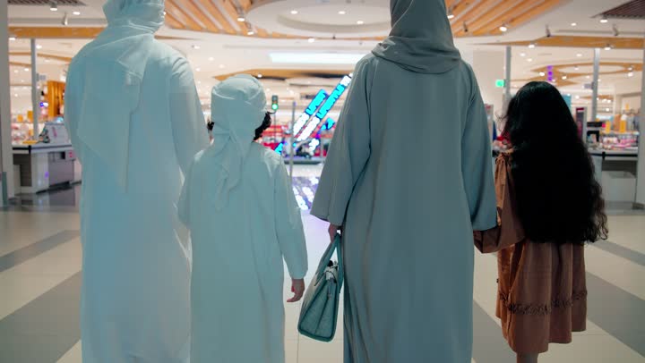 تصوير من الخلف لأب وأم عربيان خليجيان اماراتيان يرتديان الزي التقليدي يسيران برفقة ابنائهم داخل مركز تسوق عصري، قضاء وقت عائلي ممتع أثناء التسوق، مفهوم الأسرة والتسوق في المولات الحديثة