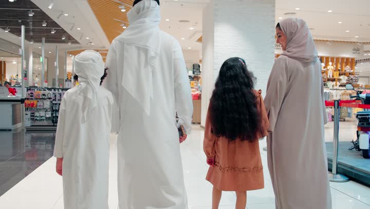 قضاء وقت عائلي ممتع أثناء التسوق، تصوير من الخلف لأب وأم عربيان خليجيان اماراتيان يرتديان الزي التقليدي يسيران برفقة ابنائهم داخل مركز تسوق عصري، مفهوم الأسرة والتسوق في المولات الحديثة