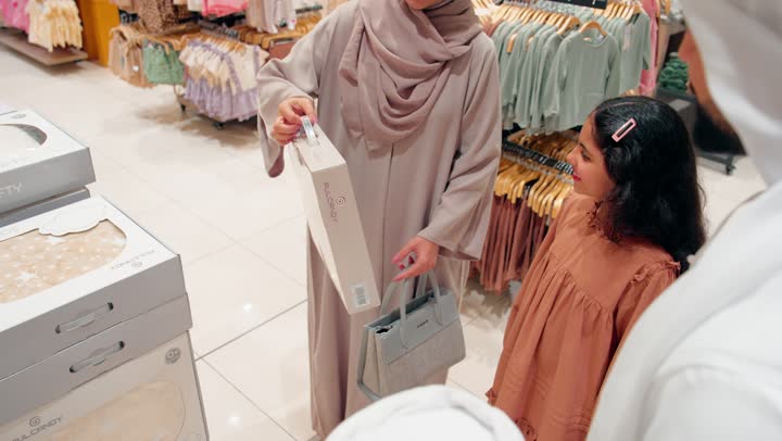 اختيار منتجات العناية بالمواليد، عائلة عربية خليجية اماراتية ترتدي الزي التقليدي داخل مركز تسوق تتفقد بطانية أطفال حديثي الولادة، مفهوم التسوق العائلي والاستعداد للطفل