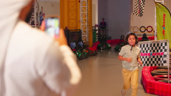 التقاط صور تذكارية عبر الهاتف المحمول ، قضاء وقت ممتع مع العائلة ، رجل عربي خليجي اماراتي يرتدي الكندورة و الغتره يقوم بتصوير ابنه في مدينه الملاهي ، تعابير الموافقة و القبول، ايماءات السعادة و السرور