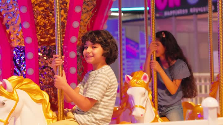 A Gulf Arab Emirati father and mother wearing traditional attire are watching their children while riding the carousel, enjoying happy family moments in an entertainment center, a young Arab girl looks on with gestures of happiness and joy.