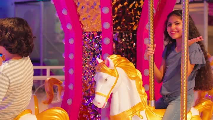 لحظات عائلية سعيدة في مركز ترفيهي، أب وأم عربيان خليجيان اماراتيان يرتدي الزي التقليدي يشاهدان ابنائهم أثناء ركوب لعبة الدوامة، فتاة عربية تلوح بيدها بايماءات السعادة و السرور