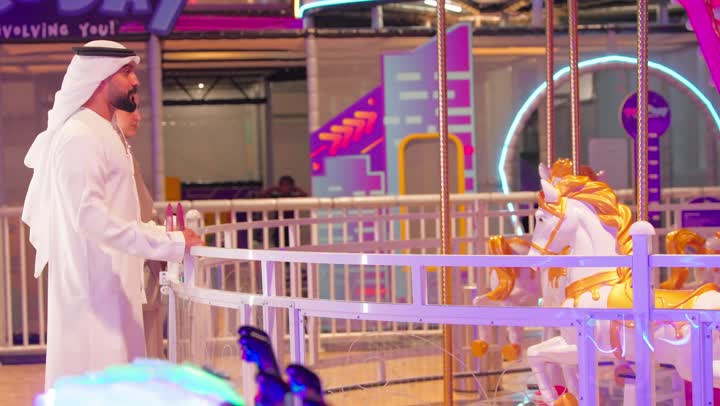 مفهوم الترابط الأسري، أب عربي خليجي اماراتي يرتدي الكندورة و الغتره  يشجع ابنه بالتصفيق أثناء ركوبه لعبة الخيل الدوارة برفقة والدته داخل الملاهي ، ايماءات السعادة و السرور، قضاء وقت عائلي ممتع