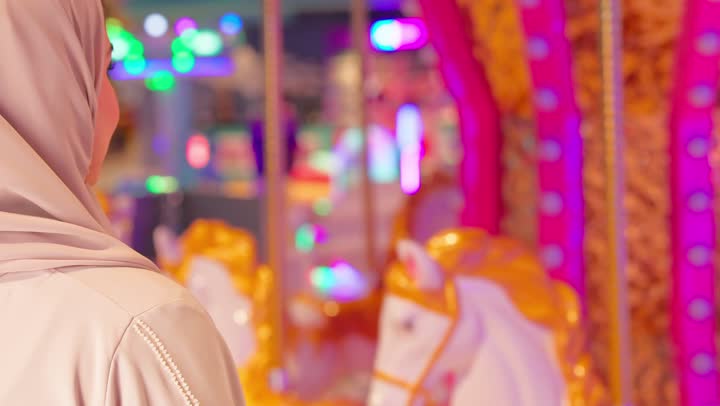 أجواء ترفيهية مبهجة داخل مدينة الألعاب، تصوير من الخلف لسيدة عربية خليجيه اماراتية محجبة ترتدي العباءة تقف بجوار لعبة الخيل الدوارة وتلوح بيديها