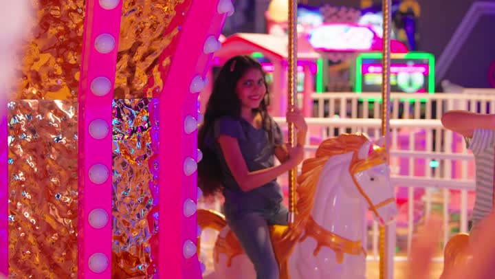 مفهوم الترفيه والتسلية، بنت عربية خليجيه اماراتية ترتدي الزي الكاجوال تستمتع بتجربة لعبة الخيل الدوارة وتلوح بيدها بابتسامة، ايماءات السعادة والسرور، قضاء وقت ممتع في مدينة الألعاب