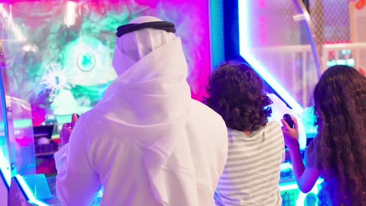تمضية أوقات من المرح مع العائلة، نشاط ترفيهي للأطفال، تصوير من الخلف لعائلة عربية خليجية اماراتية ترتدي الزي التقليدي تستمتع بالمشاركة الجماعية في لعبة صيد الأسماك الإلكترونية داخل صالة ألعاب، ايماءات التفاعل و الاندماج