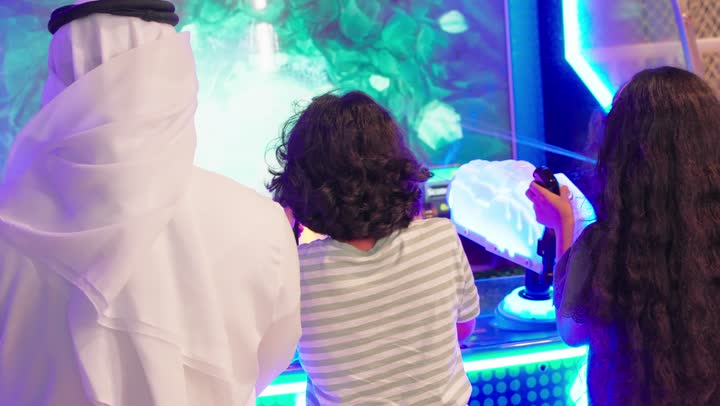 الاستمتاع بلحظات سعيدة مع العائلة، نشاط ترفيهي للأطفال، تصوير من الخلف لعائلة عربية خليجية اماراتية ترتدي الزي التقليدي تستمتع بالمشاركة الجماعية في لعبة صيد الأسماك الإلكترونية داخل صالة ألعاب، ايماءات التفاعل و الاندماج