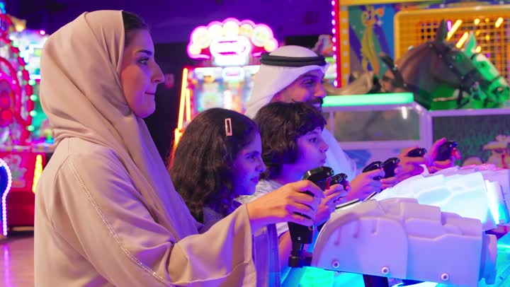 Spending quality time with family members, a recreational activity for children, an Emirati Gulf Arab family dressed in traditional attire enjoying group participation in an electronic fishing game inside an arcade, gestures of interaction and integration, the concept of family entertainment.
