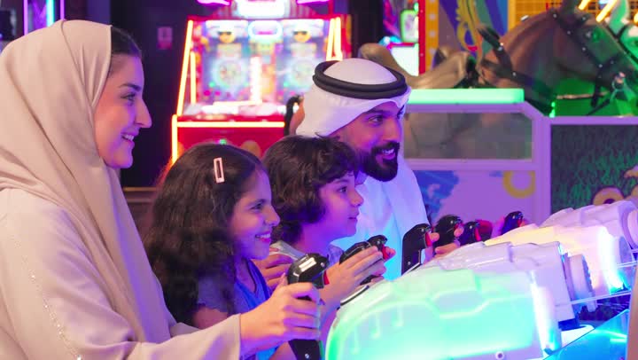 مشاركة العائلة أوقات مليئة بالمتعة، نشاط ترفيهي للأطفال، عائلة عربية خليجية اماراتية ترتدي الزي التقليدي تستمتع بالمشاركة الجماعية في لعبة صيد الأسماك الإلكترونية داخل صالة ألعاب، ايماءات التفاعل و الاندماج، مفهوم الترفية العائلي