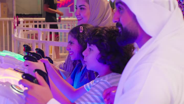 Enjoying special moments with the family, a recreational activity for children with family participation, an Arab Gulf Emirati family dressed in traditional attire enjoying group participation in an electronic fishing game inside an arcade, gestures of interaction and integration, the concept of family entertainment.