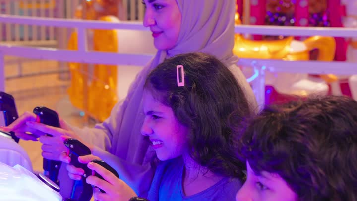 قضاء أوقات ممتعة مع العائلة، نشاط ترفيهي للأطفال بمشاركة الاسرة، عائلة عربية خليجية اماراتية ترتدي الزي التقليدي تستمتع بالمشاركة الجماعية في لعبة صيد الأسماك الإلكترونية داخل صالة ألعاب، ايماءات التفاعل و الاندماج، مفهوم الترفية العائلي