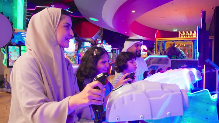 نشاط عائلي ترفيهي للأطفال، عائلة عربية خليجية اماراتية ترتدي الزي التقليدي تستمتع بالمشاركة الجماعية في لعبة صيد الأسماك الإلكترونية داخل صالة ألعاب، ايماءات التفاعل و الاندماج، مفهوم الترفية العائلي