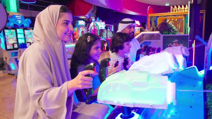 عائلة عربية خليجية اماراتية ترتدي الزي التقليدي تستمتع بالمشاركة الجماعية في لعبة صيد الأسماك الإلكترونية داخل صالة ألعاب، ايماءات التفاعل و الاندماج، مفهوم الترفية العائلي
