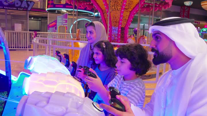 ايماءات التفاعل و الاندماج، مفهوم الترفية العائلي، عائلة عربية خليجية اماراتية ترتدي الزي التقليدي تستمتع بالمشاركة الجماعية في لعبة صيد الأسماك الإلكترونية داخل صالة ألعاب
