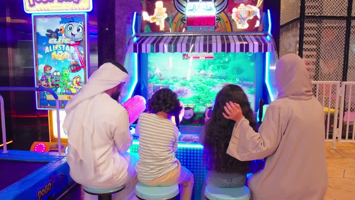 مفهوم الترفية العائلي، تصوير من الخلف لعائلة عربية خليجية اماراتية ترتدي الزي التقليدي تستمتع بالمشاركة الجماعية في لعبة صيد الأسماك الإلكترونية داخل صالة ألعاب