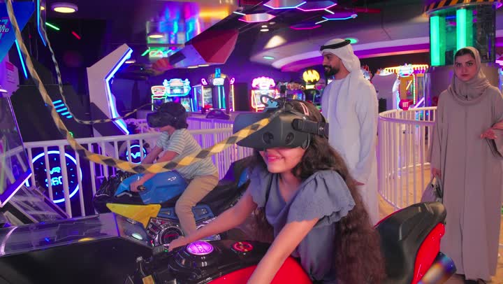 الانشطة الترفيهية الممتعه،  قضاء أوقات ممتعة مع العائلة، أب عربي خليجي اماراتي يرتدي الكندورة و الغتره يشجع طفليه أثناء اللعب في لعبة سباق الدراجات النارية بالواقع الافتراضي داخل صالة ألعاب ترفيهية حديث، الاستمتاع بتجربة لعب مميزة وآمنة