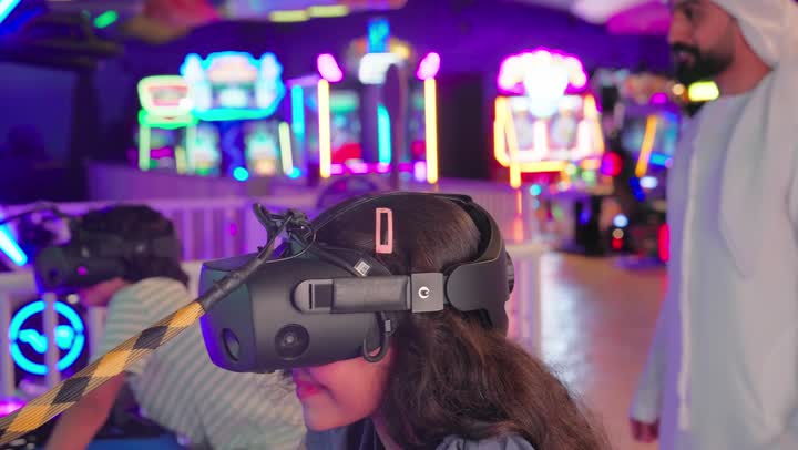 The concept of entertainment and leisure spending enjoyable time with the family. An Emirati Gulf Arab father, wearing a kandura and ghutra, encourages his children while they play a virtual reality motorcycle racing game inside a modern entertainment center, enjoying a unique and safe gaming experience.