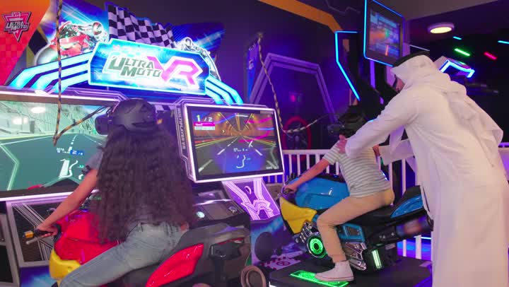Spending quality time with the family, an Emirati Gulf Arab father, wearing a kandura and ghutra, encourages his children as they play a virtual reality motorcycle racing game inside a modern entertainment center, enjoying a unique and safe gaming experience.